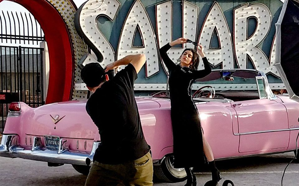 A private photoshoot in with the Sahara sign in the background, with a woman posing in front of a classic pink Cadillac.