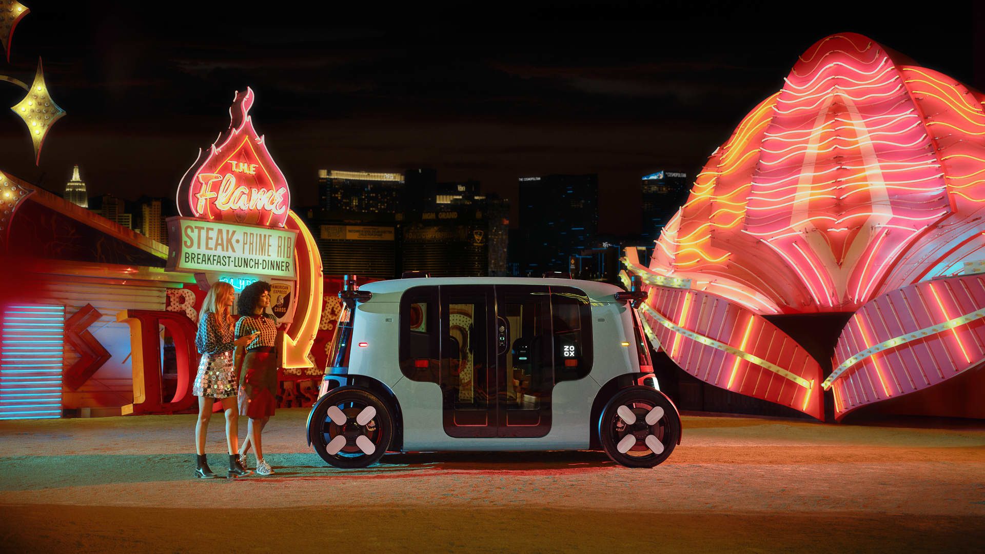 Two people next to a Zoox taxi car at the Neon Boneyard with the Las Vegas city in the background