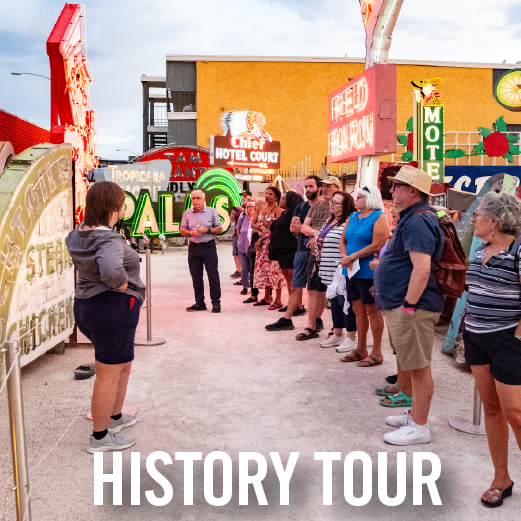 A tour guide speaks to a group of visitors standing outdoors among colorful vintage neon signs during a daytime history tour. The group listens attentively as classic Las Vegas-style signage surrounds them.