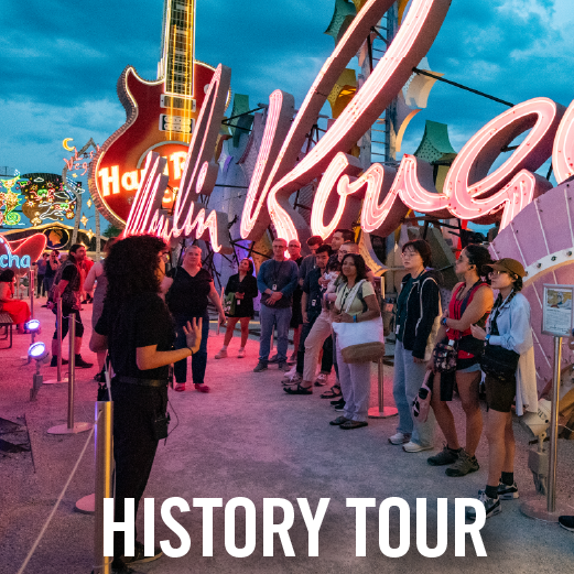 A guide addresses a group of people at night beneath glowing neon signs, including a large pink guitar sign. The visitors stand along a walkway, illuminated by bright, historic neon lights during an evening history tour.