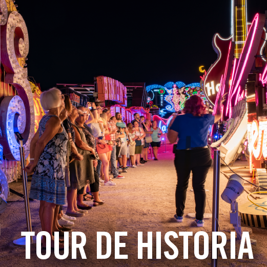 Un guía se dirige a un grupo de personas por la noche, bajo letreros de neón brillantes, incluido un gran letrero con forma de guitarra rosa. Los visitantes están de pie a lo largo de un pasillo, iluminados por luces de neón históricas y resplandecientes durante un recorrido histórico nocturno.