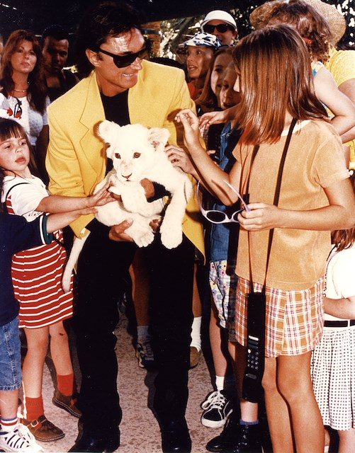 Photograph of Roy Horn standing surrounded by children holding a white tiger.